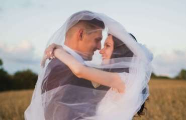 A married couple embracing in a field, the bride's veil getting blown over by wind over the head of the groom